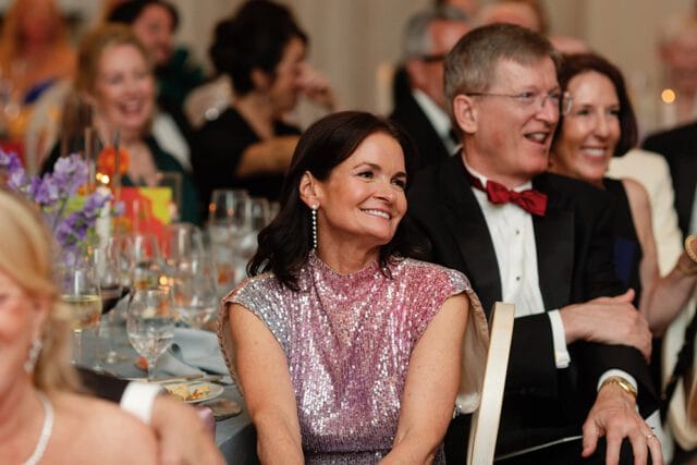 Woman in a sparkly pink and silver dress smiles while seated at a formal dinner table next to a man in a tuxedo with a red bow tie.