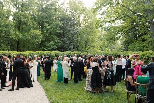 A large group of people in formal attire mingle on a grassy lawn surrounded by lush trees and manicured hedges during an outdoor event.
