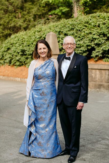 Couple posing outdoors, woman in a blue patterned gown and white shawl, man in a tuxedo.