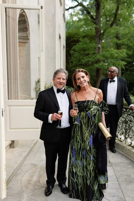 Man in a tuxedo holding a champagne flute and woman in a floral dress holding a champagne flute, with another man in a tuxedo in the background.