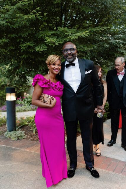 A Black couple smiles at the camera, the woman in a bright pink off-the-shoulder gown with ruffled trim and the man in a black tuxedo with a bow tie and glasses.