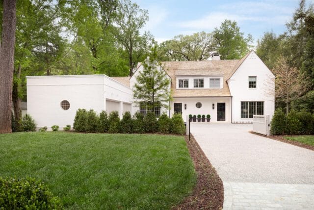 White brick house with a light wood shingle roof, black-framed windows, and a circular window on the garage. A gravel driveway leads to the entrance, flanked by manicured hedges and a lush green lawn.