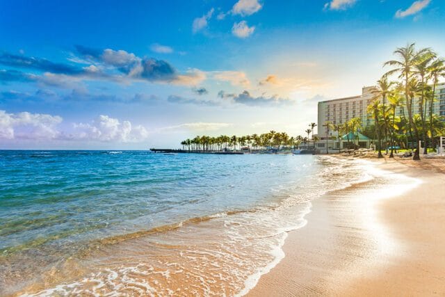 Beach with turquoise water, golden sand, palm trees, and a large hotel building under a blue sky with scattered clouds.