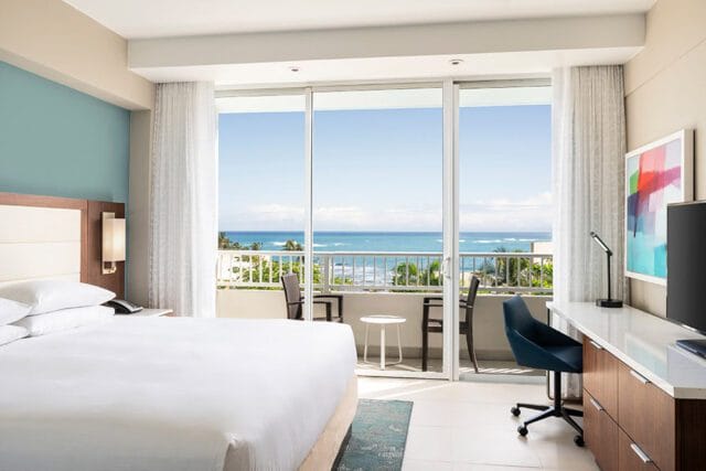 Bedroom with a king-size bed, light blue accent wall, and a desk with a TV, overlooking the ocean from a private balcony.