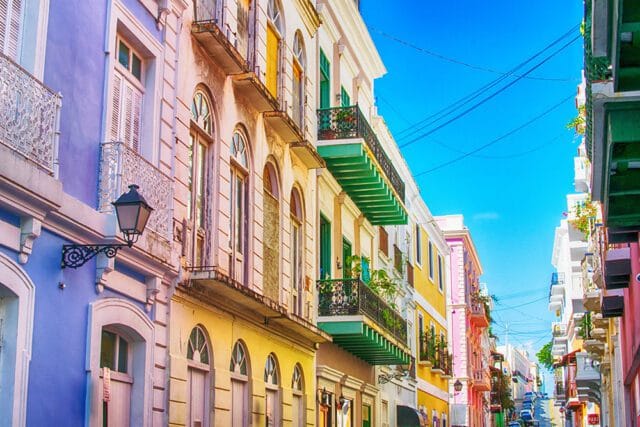 Row of colorful colonial buildings with wrought iron balconies and arched windows on a narrow street under a bright blue sky.