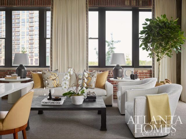 Living room with exposed brick walls, cream sofa with patterned pillows, two white armchairs with a yellow throw, and a dark coffee table with books and a plant.
