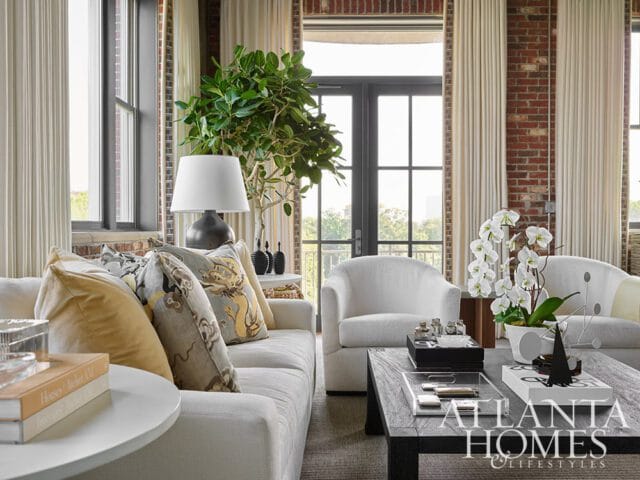 Living room with white sofa, white armchairs, dark wood coffee table, and exposed brick walls. A large plant and white orchids add greenery.