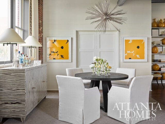 Dining area with a dark wood table, four white slipcovered chairs, a striped sideboard with a bar setup, and abstract yellow artwork on a shiplap wall.