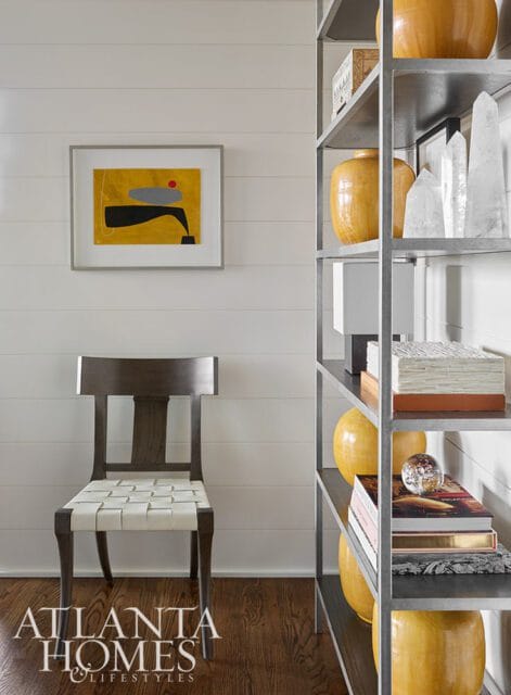 Modern living room with shiplap walls, a dark wood chair with white woven seat, abstract art, and a metal shelving unit displaying yellow vases and crystal objects.