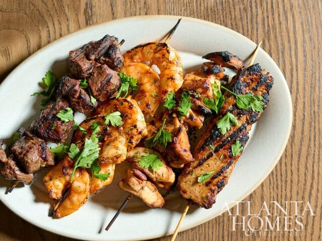 Grilled skewers of steak, shrimp, and chicken garnished with parsley, served on a light-colored oval plate with a wood-grain background.
