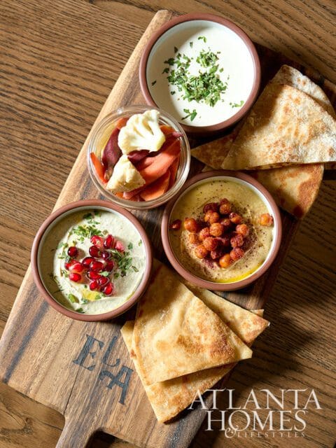 Hummus platter with pita bread, pickled vegetables, and dips including one topped with pomegranate seeds and another with chickpeas.