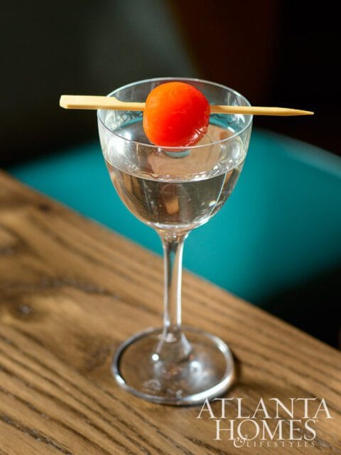 Clear cocktail in a coupe glass garnished with a cherry on a wooden skewer, with the Atlanta Homes & Lifestyles logo in the lower right corner.