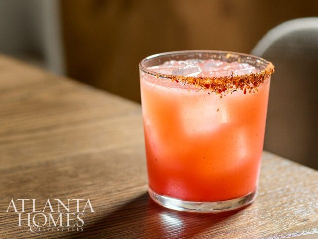 Glass of pink cocktail with ice and a spiced rim, resting on a wooden table. Atlanta Homes & Lifestyles logo in the lower left corner.