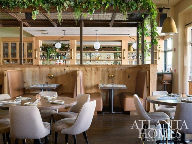Restaurant interior with wood booths, round tables, and chairs. A bar area with shelves stocked with bottles is visible in the background, along with hanging plants and pendant lights.