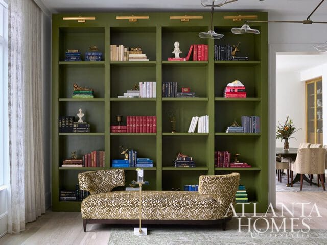 Living room with a large olive green built-in bookshelf filled with books and decorative objects. A patterned chaise lounge sits in front of the bookshelf.