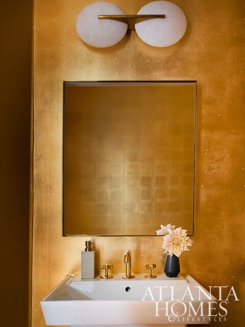 Powder room with gold textured walls, a square mirror, brass fixtures, a white sink, and a modern wall sconce with two white globes.