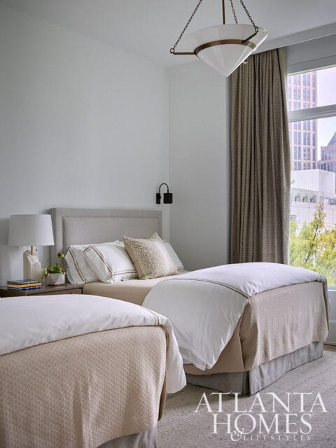 Bedroom with two twin beds, neutral bedding, upholstered headboards, and a large window with city view. Atlanta Homes & Lifestyles logo in the corner.