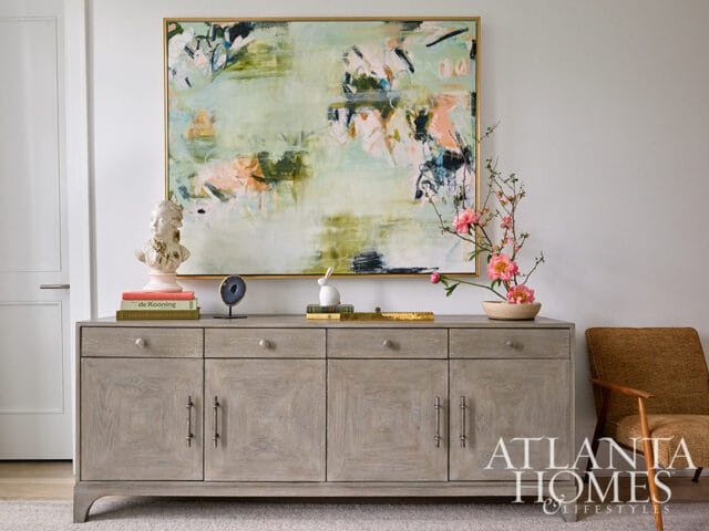 Light gray wood sideboard with brass hardware topped with books, a bust, and a floral arrangement, beneath a large abstract painting.