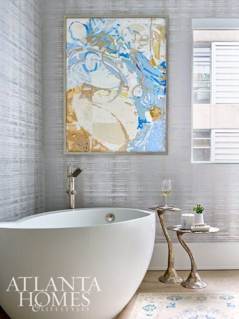Modern bathroom with a freestanding white bathtub, abstract art on a textured silver wall, and two ornate side tables.