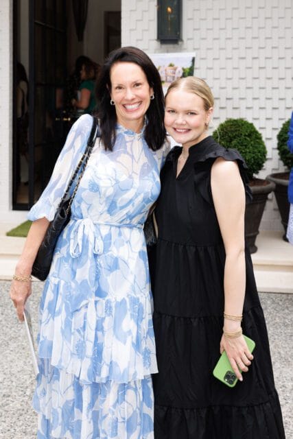 Two women smiling at the camera outside a white brick house. The woman on the left wears a blue and white floral dress and the woman on the right wears a black dress.