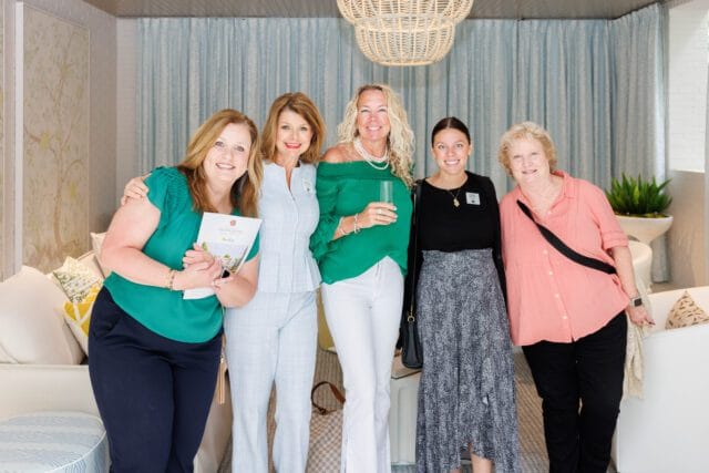 Five women pose for a photo in a room with light blue patterned curtains, a large woven pendant light, and white furniture. One woman holds a brochure for Southeastern Showhouse.