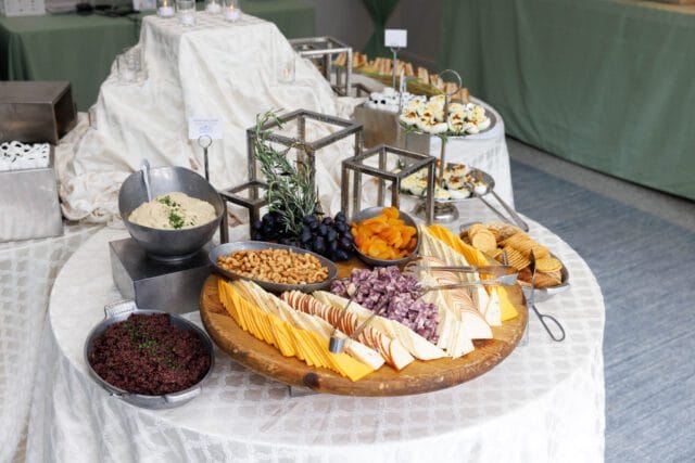 Buffet table with a round wooden platter of sliced cheeses, crackers, grapes, dried apricots, and nuts. Other dishes include deviled eggs, hummus, and a dark spread.