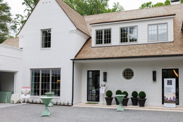 White brick house with cedar shake roof, large windows, and a black front door. Outdoor cocktail tables and potted topiaries are set up in front.