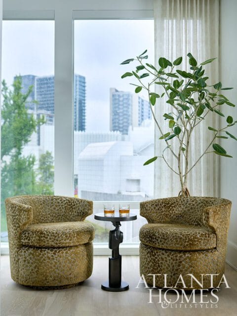 Two upholstered swivel chairs with a leopard-like pattern flank a small dark metal table holding two glasses of amber liquid. A tall plant stands behind the table.