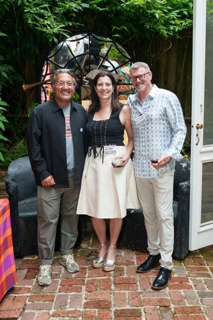 Three people posing for a photo in a garden setting. The man on the left wears a black jacket over a gray t-shirt and khaki pants. The woman in the center wears a black corset top and a cream-colored skirt. The man on the right wears a patterned blue and white shirt and light-colored pants.