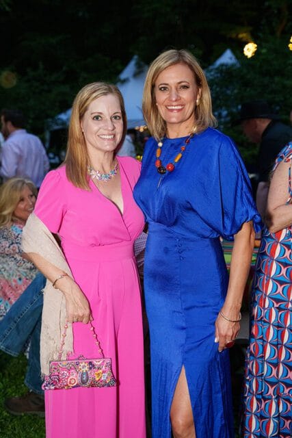 Two women smiling at an outdoor event. The woman on the left wears a bright pink jumpsuit and a light shawl, holding a colorful beaded clutch. The woman on the right wears a royal blue dress and a large beaded necklace.