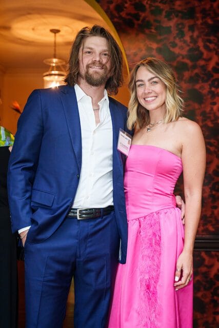 Man in a blue suit and white shirt stands next to a woman in a pink strapless dress with feathers.