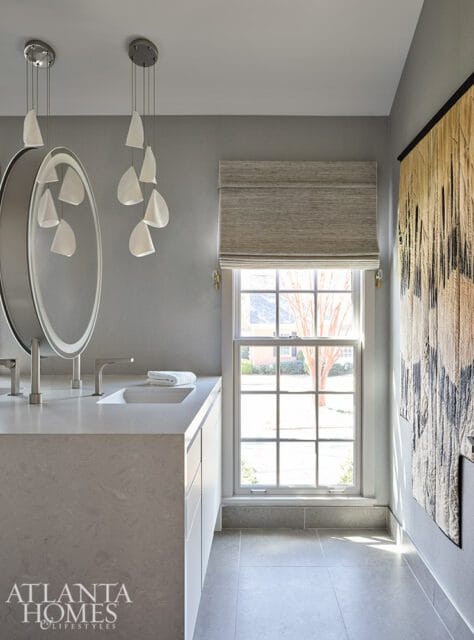 Gray bathroom with a floating vanity, oval mirror with integrated lighting, modern pendant lights, and a large window with a Roman shade.