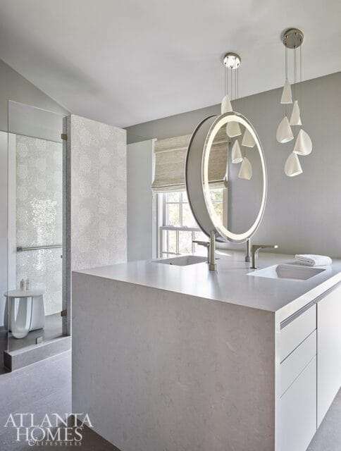 Modern bathroom with a large gray stone vanity featuring two sinks, a circular backlit mirror, and contemporary pendant lighting. A glass-enclosed shower is visible to the left.