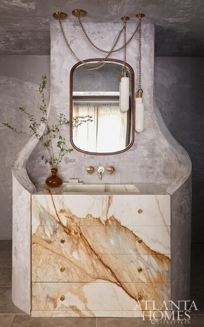 Bathroom vanity with a marble countertop and drawers featuring dramatic brown veining, a rectangular sink, and brass fixtures. Above, a mirror is framed by a dark wood border, and two pendant lights hang from chains.