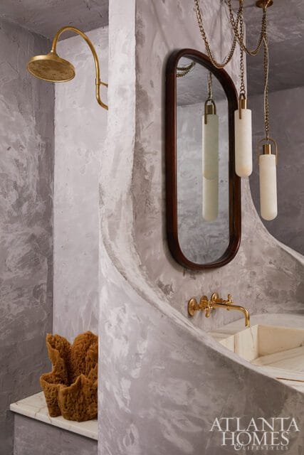 Bathroom with textured gray walls, brass showerhead, oval mirror with dark wood frame, and a curved vanity with a brass faucet.