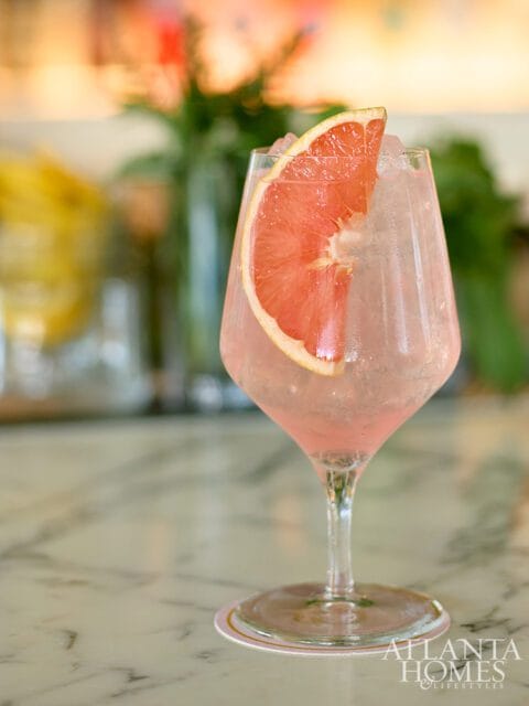 Pink cocktail in a wine glass with ice and a grapefruit slice garnish, on a marble countertop with a coaster.