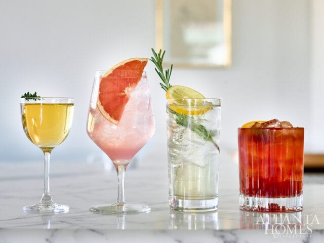 Four cocktails on a marble countertop: a yellow drink with thyme, a pink drink with grapefruit and rosemary, a clear drink with lemon and rosemary, and a red drink with orange slice.