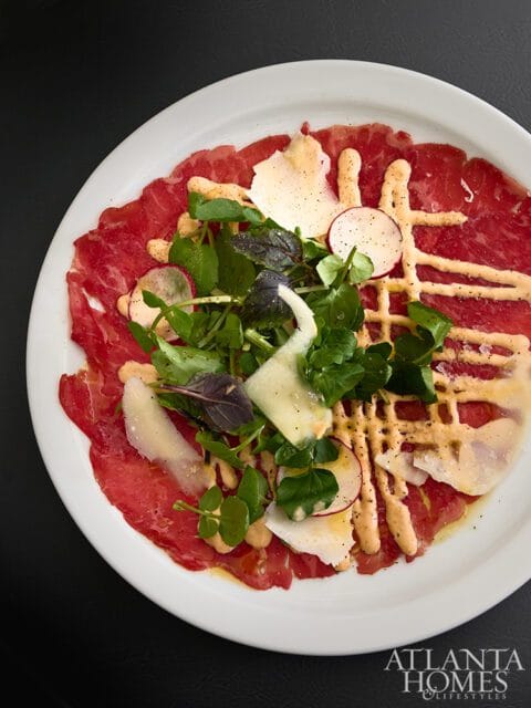 Thinly sliced beef carpaccio topped with arugula, shaved parmesan, radish slices, and a drizzle of creamy sauce on a white plate.