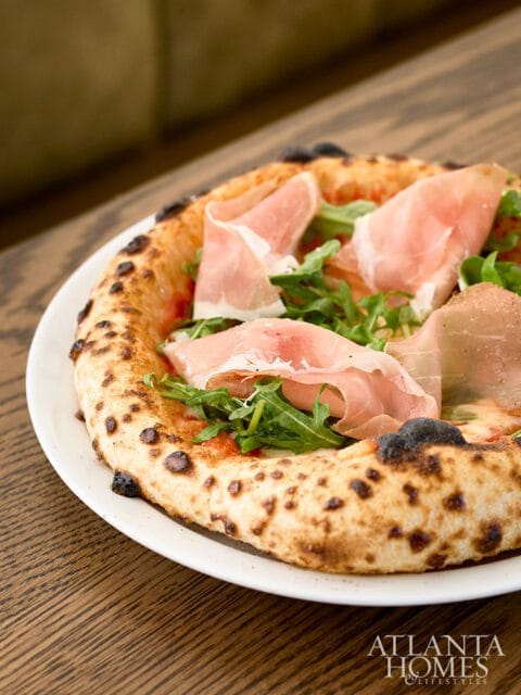 Neapolitan pizza with prosciutto and arugula on a white plate, with a dark wood table surface and a blurred background.