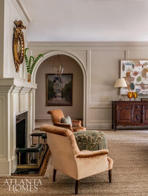 Living room with a fireplace, two velvet armchairs in peach and rust tones, a green patterned ottoman, and a dark wood cabinet with abstract art above.