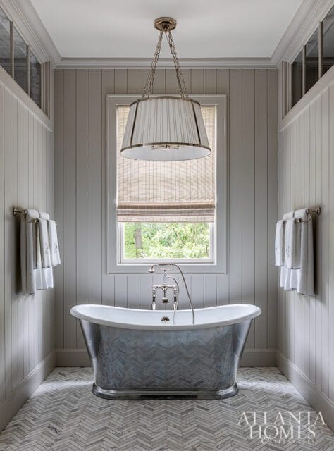 Gray paneled bathroom with a freestanding clawfoot tub featuring a metallic herringbone pattern, a large pleated drum pendant light, and a herringbone tile floor.