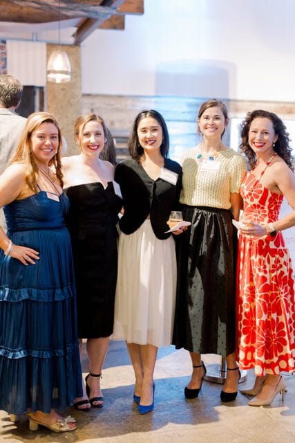 Five women posing for a photo at an event. From left to right: a woman in a blue tiered dress, a woman in a black strapless dress, a woman in a black jacket and white skirt, a woman in a yellow textured top and black skirt, and a woman in a red floral dress.