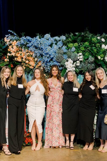 Seven women stand in front of a floral backdrop featuring blue hydrangeas, orange flowers, and greenery. The woman in the center wears a pink floral dress, while others wear black or white attire.