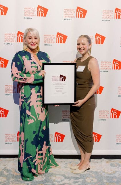 Two women stand in front of a backdrop with the American Society of Interior Designers Georgia logo. The woman on the left wears a green, pink, and navy floral dress and holds a framed award. The woman on the right wears a brown sleeveless dress and has a name tag.