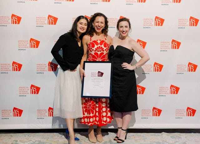Three women pose for a photo in front of a backdrop with the American Society of Interior Designers Georgia logo repeated. The woman in the center holds a framed award.