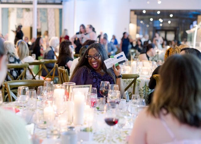 Woman in glasses smiles and holds up a card at a formal dinner event with candles and wine glasses on the table.