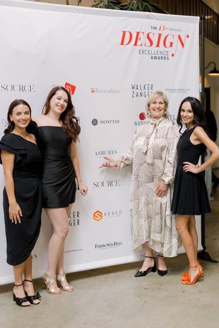 Four women pose in front of a white backdrop with "The 43rd Annual Design Excellence Awards" logo and sponsor names. The women wear black and patterned dresses.