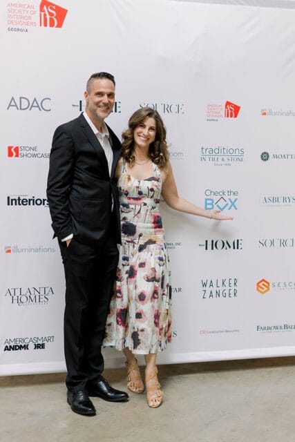 Man in a black suit and woman in a floral dress pose in front of a white backdrop with sponsor logos including ADAC, Stone Showcase, Atlanta Homes & Lifestyles, and Traditions in Tile & Stone.
