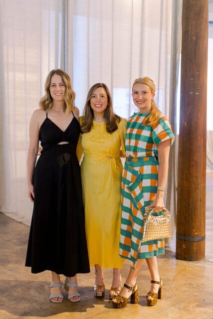 Three women standing together, the woman on the left wears a black dress, the middle woman wears a yellow dress, and the woman on the right wears a green and orange checkered dress and holds a gold purse.
