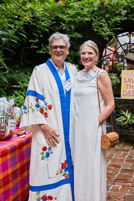Man wearing a white robe with blue trim and floral embroidery, and a woman in a white dress, posing outdoors with lush greenery in the background.